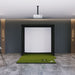 Indoor golf simulator setup with green mat and white screen on a gray floor.